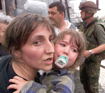 Woman clinging to crying child while other people, including an American soldier, contemplate a scene of carnage.